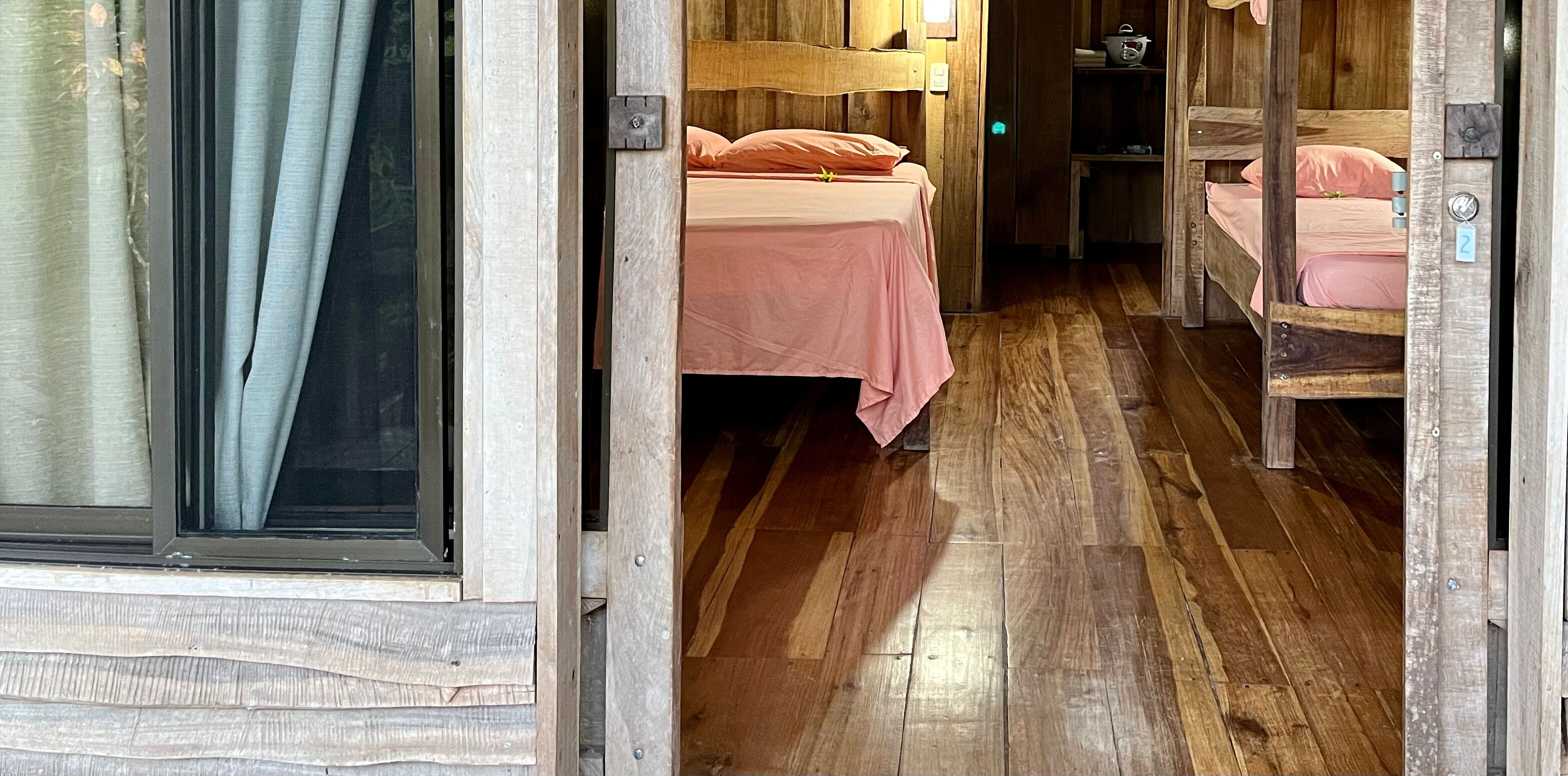 A wooden interior with two beds, a wooden floor, and a window with curtains.