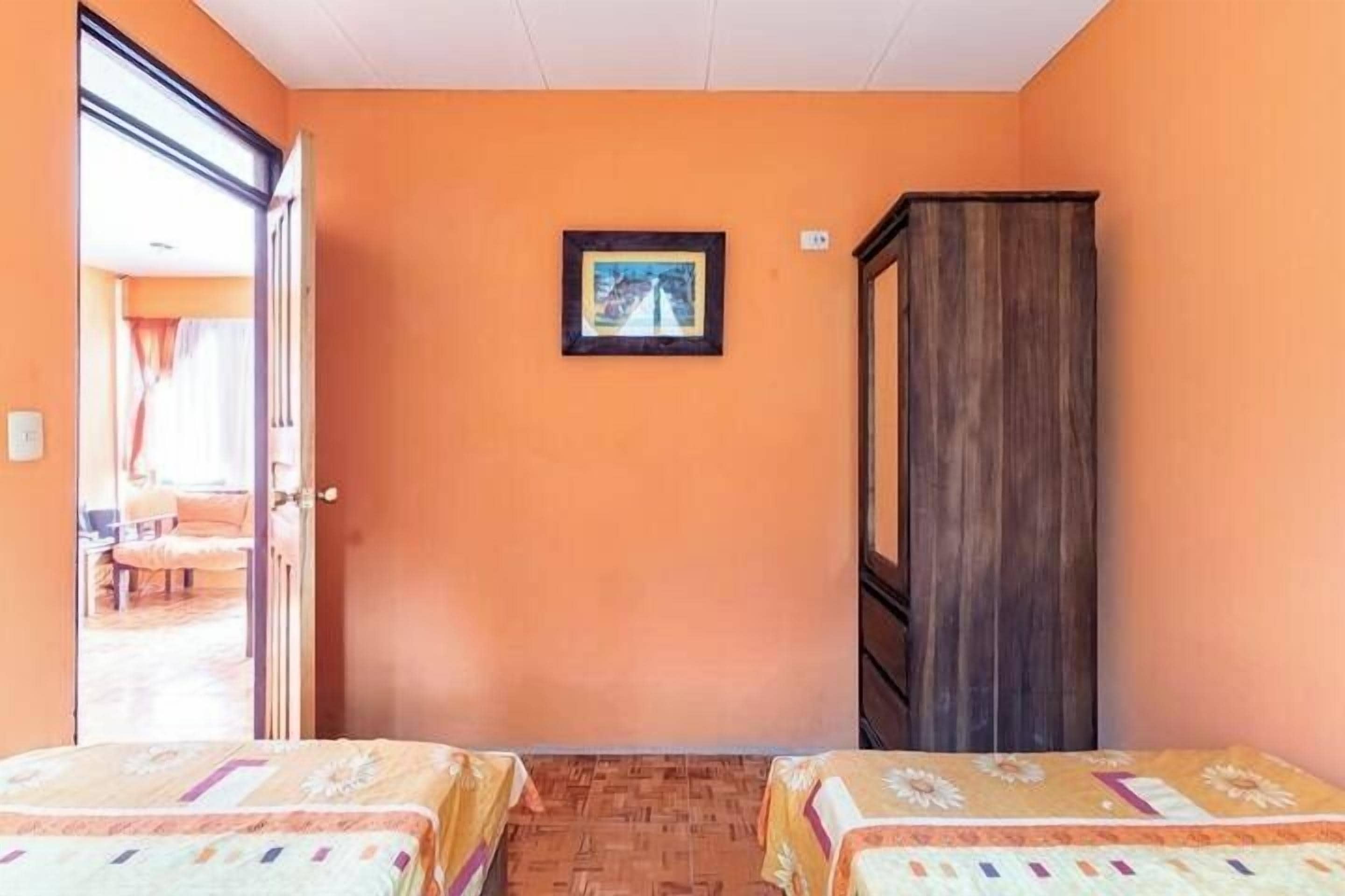 Una habitación con dos camas, un armario de madera, un cuadro enmarcado en la pared y una puerta abierta que da a otra habitación.