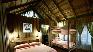 A room with wooden walls and a sloped ceiling, featuring bunk beds, a desk with a chair, and a window with curtains.