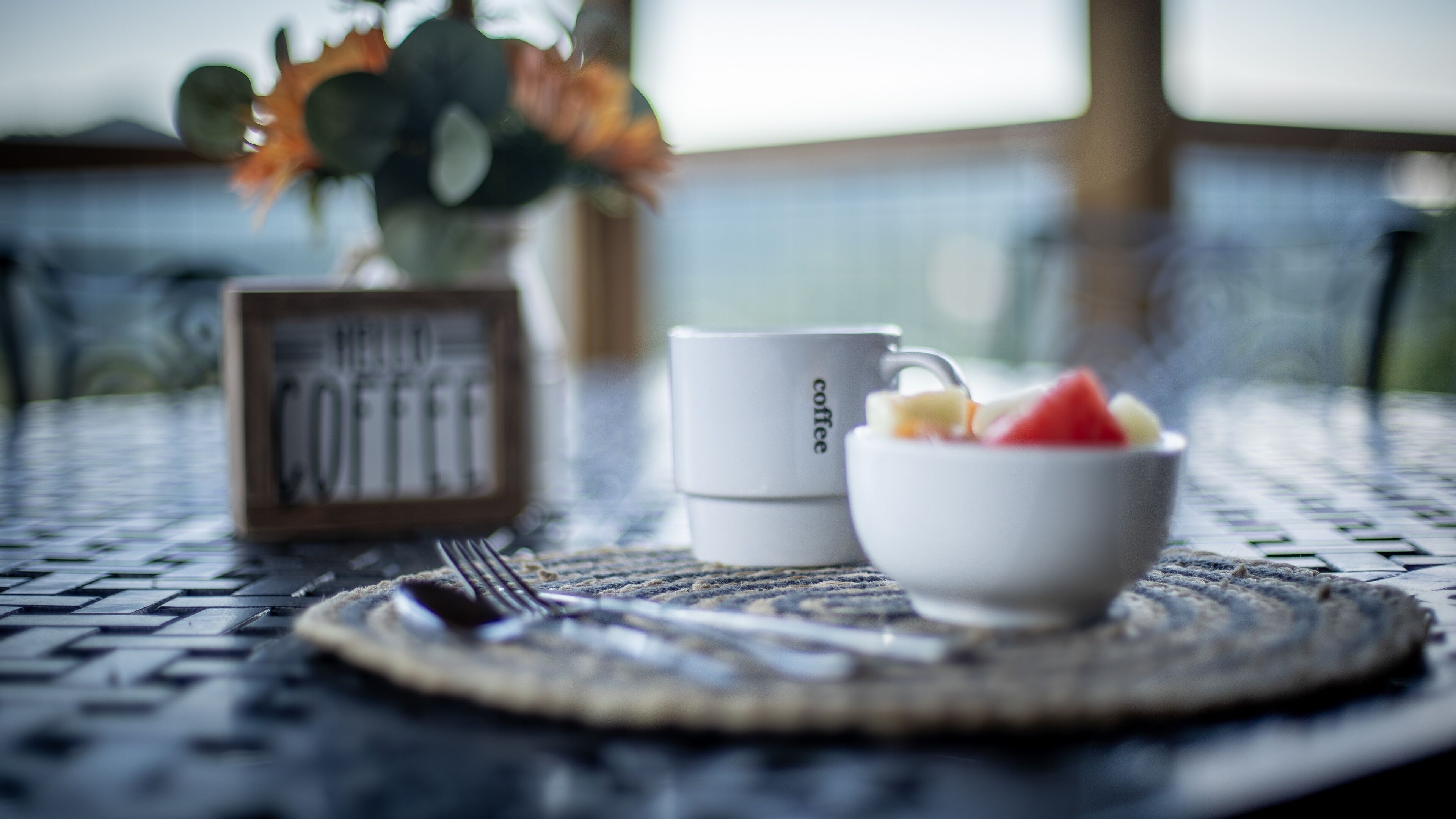 A breakfast setting with a coffee mug, a bowl of fruit, and a framed picture on a table.