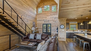 A spacious living room with a brown leather sofa, a coffee table, and a staircase leading to an upper level. A kitchen area is visible in the background with a sign labeled "KITCHEN". - The Hidden Owl Mountain Lodge 5 Bedroom Cabin by RedAwning (Sevierville)