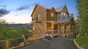A large wooden cabin with a stone extension, a covered porch, and a view of the surrounding forest. - The Hidden Owl Mountain Lodge 5 Bedroom Cabin by RedAwning (Sevierville)