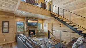 A cabin interior with a stone fireplace, leather sofas, a staircase, and a large window overlooking a mountainous landscape. - The Hidden Owl Mountain Lodge 5 Bedroom Cabin by RedAwning (Sevierville)