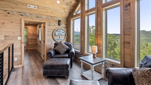 A wooden interior with a sofa, ottoman, and table near large windows overlooking a forested landscape. - Red Sky Ridge 5 Bedroom Cabin by RedAwning (Sevierville)