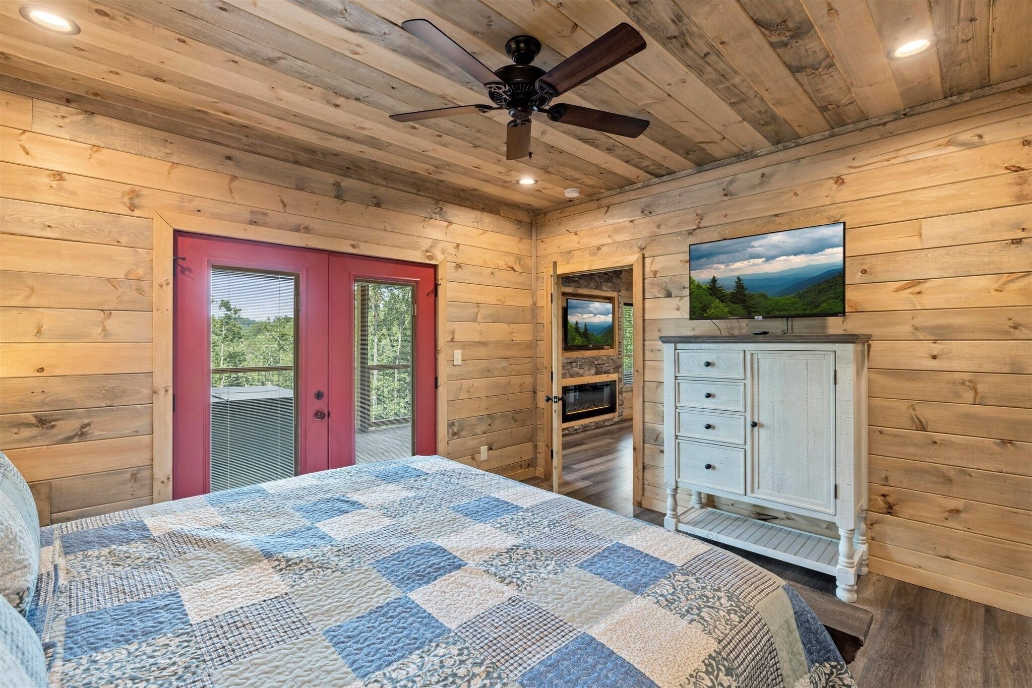 A wooden cabin bedroom with a bed, a ceiling fan, a red door, a television, and a white dresser.