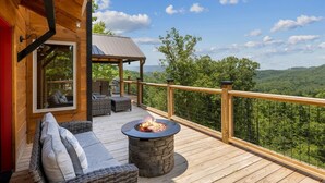 A wooden deck with a stone fire pit, outdoor seating, and a view of a forested landscape. - Red Sky Ridge 5 Bedroom Cabin by RedAwning (Sevierville)