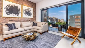 A modern living room with a sectional sofa, a glass coffee table, and a leather armchair. There are two framed artworks on the brick wall, and a large sliding glass door leading to a balcony with a view of buildings. - Perfect, freshly decorated one bedroom Loft 268! (Puerto Vallarta)