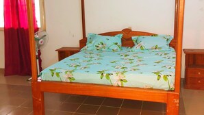 Ein Bett mit Baldachin, eine blumige Tagesdecke, ein runder Teppich auf dem Boden und ein Standventilator in der Nähe eines Fensters mit roten Vorhängen.