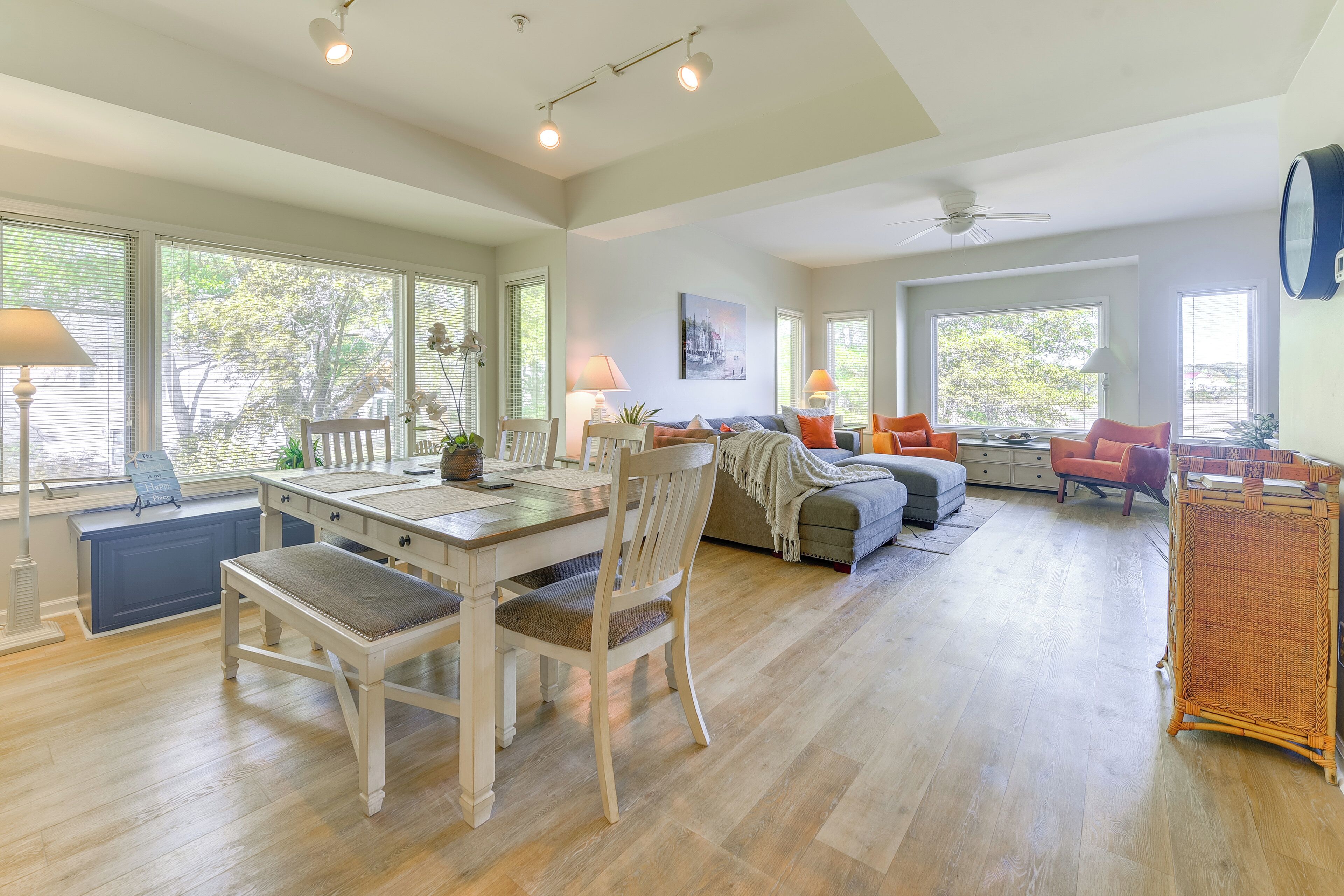 A spacious living area with a dining table, chairs, and a sofa. There are large windows providing natural light and views of the outdoors.