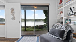 Habitación con puerta de vidrio corredera que da a vistas de un lago, sofá gris con cojines estampados, y una estantería con diversos objetos.