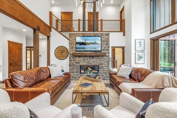 A spacious living room with leather sofas, a stone fireplace, a wooden staircase, and a large TV mounted above it. - 5 Star Luxury Crafted for Family Memories | Spa/Epic GameRm+ 1PM Check In/Out (Cle Elum)