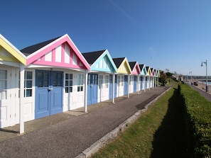 A row of colorful beach huts with white doors and windows, aligned along a paved path beside a grassy area. - The White Cottage (Weymouth)