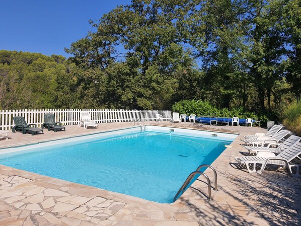 Ein rechteckiger Swimmingpool mit einer Bordüre aus Steinplatten, umgeben von weißen Liegestühlen und einem weißen Zaun.