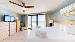 A hotel room with a bed, a television, a desk, and a view of the beach.
