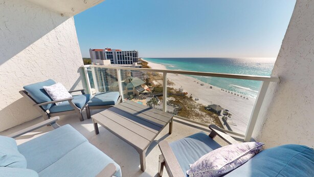 A balcony with a view of a beach and ocean. There are lounge chairs and a table.
