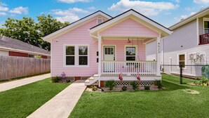 Una casa de un solo piso de color rosa con un porche delantero, un pequeño jardín frontal y una cerca de madera.