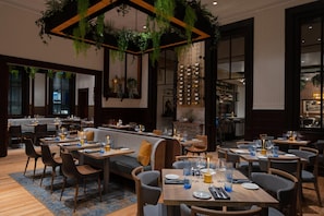 A restaurant with multiple dining tables and booths, a bar area, and hanging plants. - Hotel West & Main Conshohocken Philadelphia, Tapestry Collection (Conshohocken)