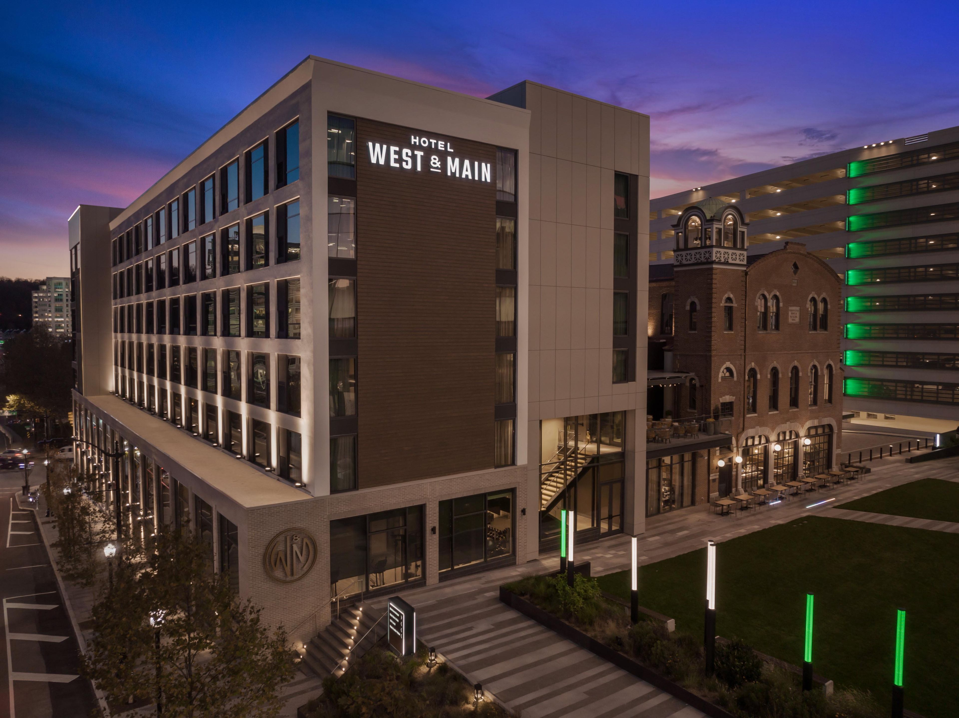 A modern hotel building with a prominent sign reading 'HOTEL WEST & MAIN' and a traditional building with green lighting in the background.