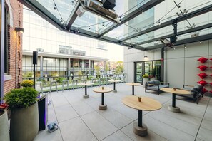 A modern outdoor seating area with round tables and chairs, a glass ceiling, and a view of a building with large windows. - Hotel West & Main Conshohocken Philadelphia, Tapestry Collection (Conshohocken)