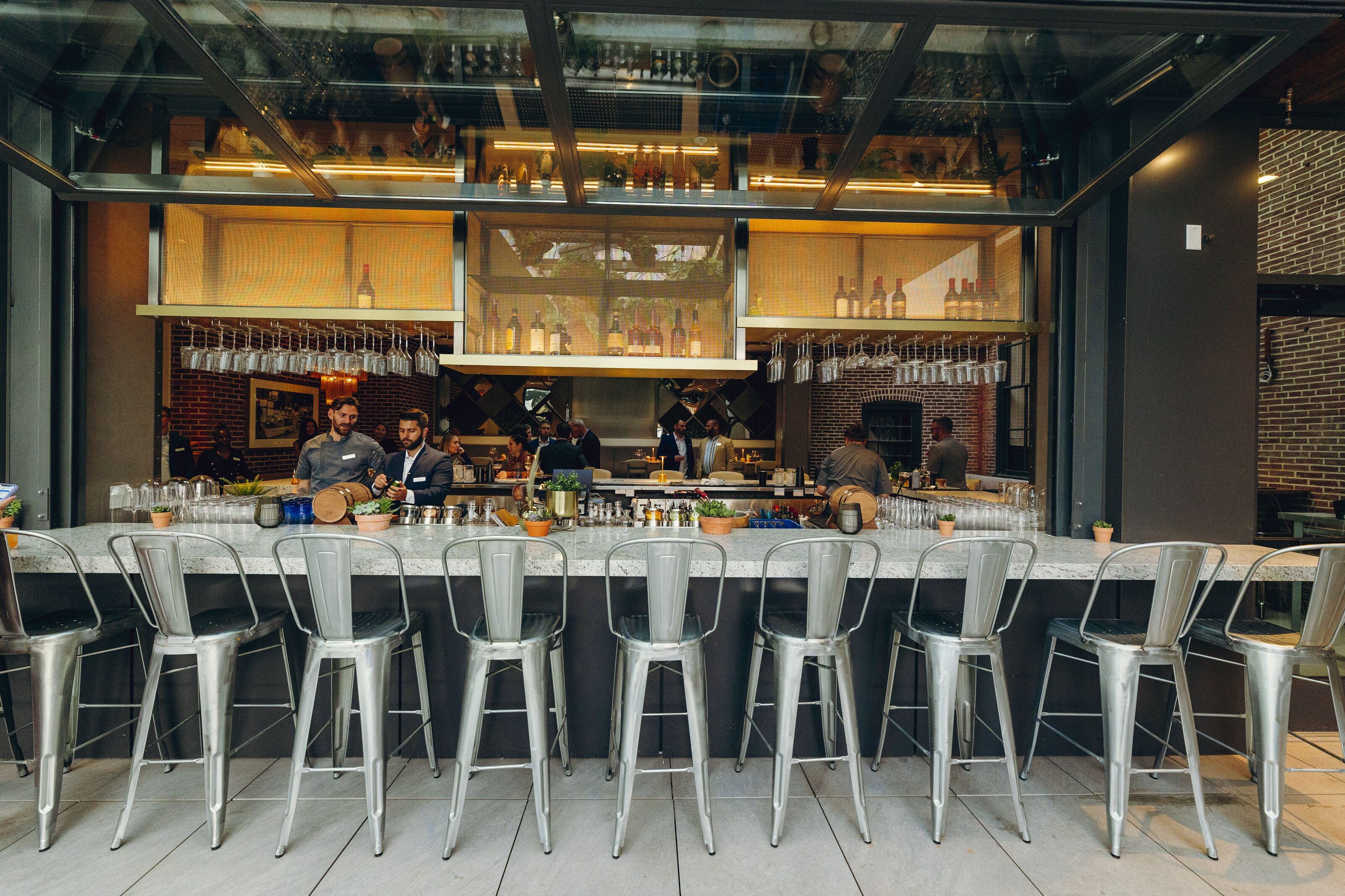 Eine Bar mit einer langen Theke, Metallstühlen und Regalen voller Flaschen und Gläsern.