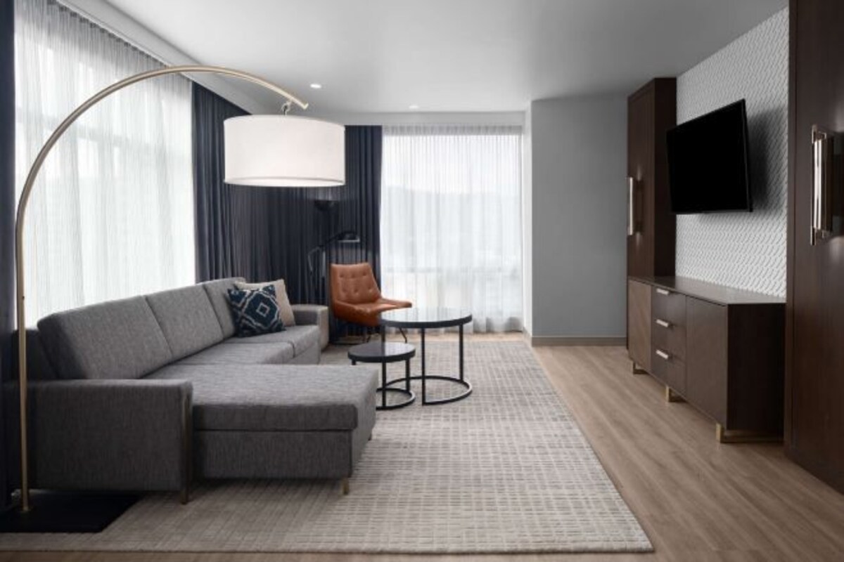 Una habitación de hotel moderna con un sofá seccional gris, un sillón de cuero marrón, una mesita redonda pequeña, una lámpara de pie, un televisor montado en una pared texturizada y suelo de madera.