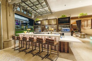 A modern bar area with a high ceiling, bar stools, a counter, and a large screen. - Hotel West & Main Conshohocken Philadelphia, Tapestry Collection (Conshohocken)