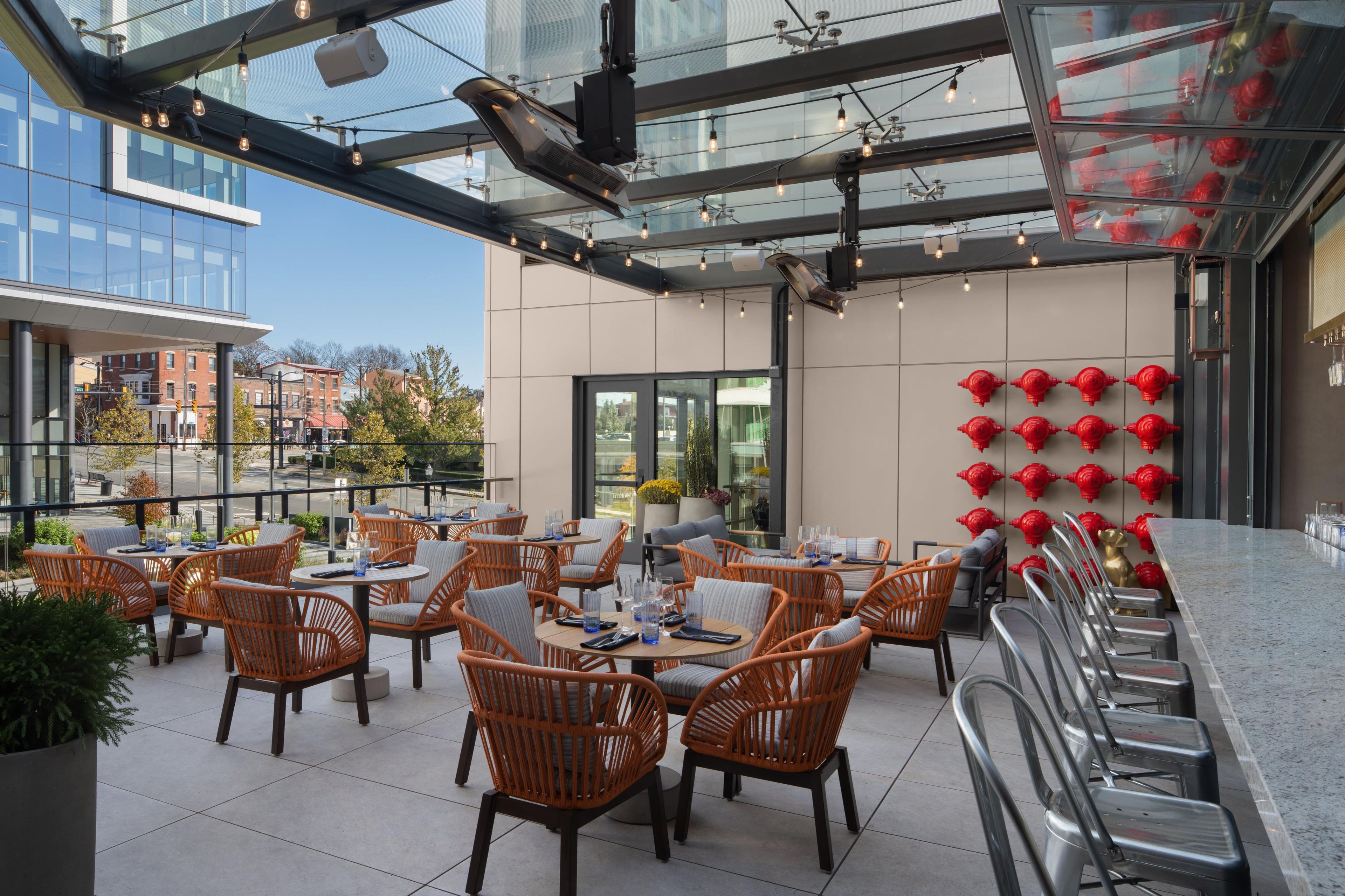 An outdoor seating area with wicker chairs and tables, a glass ceiling, and a row of red fire hydrants on the wall.