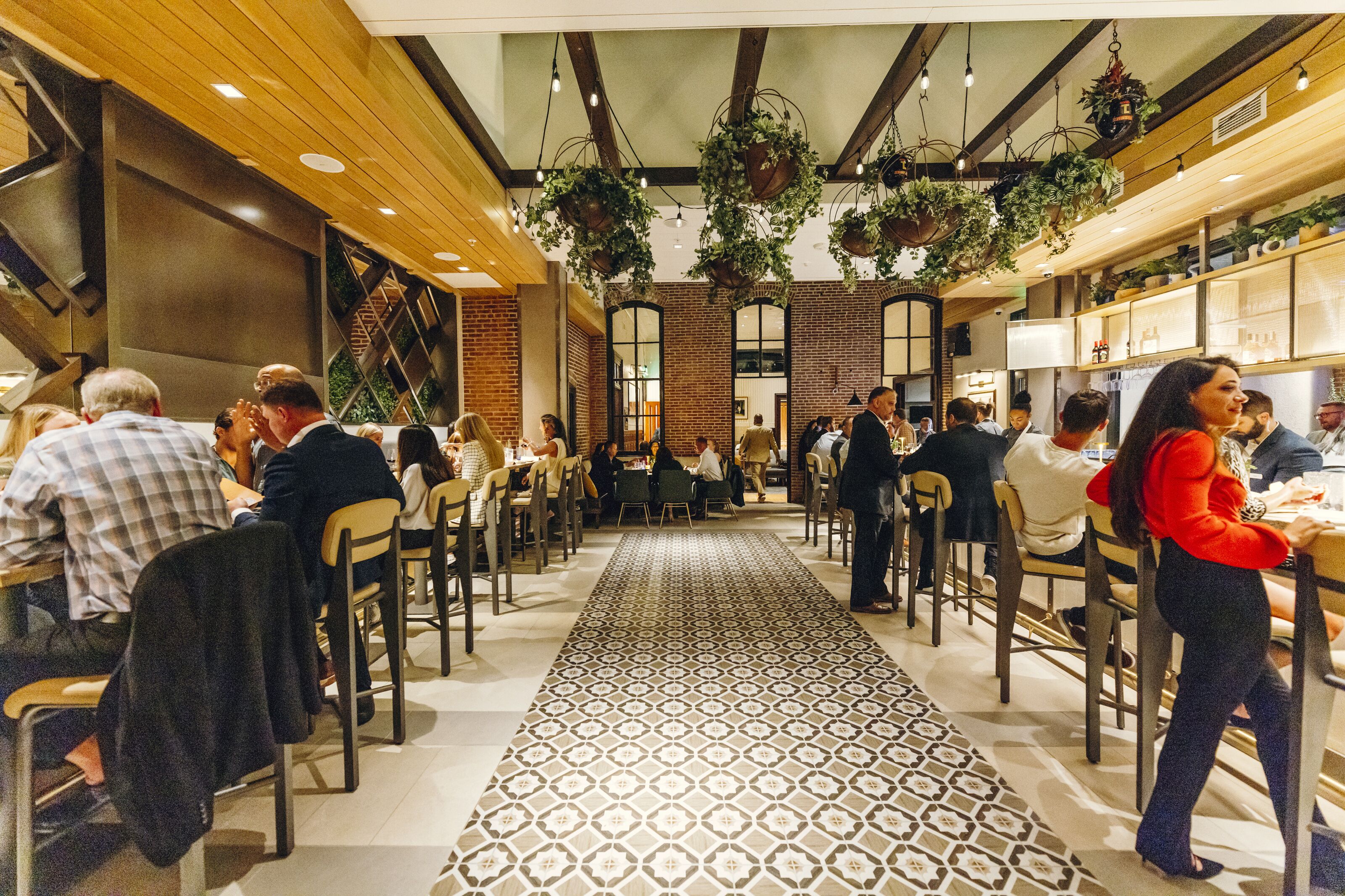 A restaurant with a patterned floor, wooden ceiling beams, hanging plants, and brick walls.