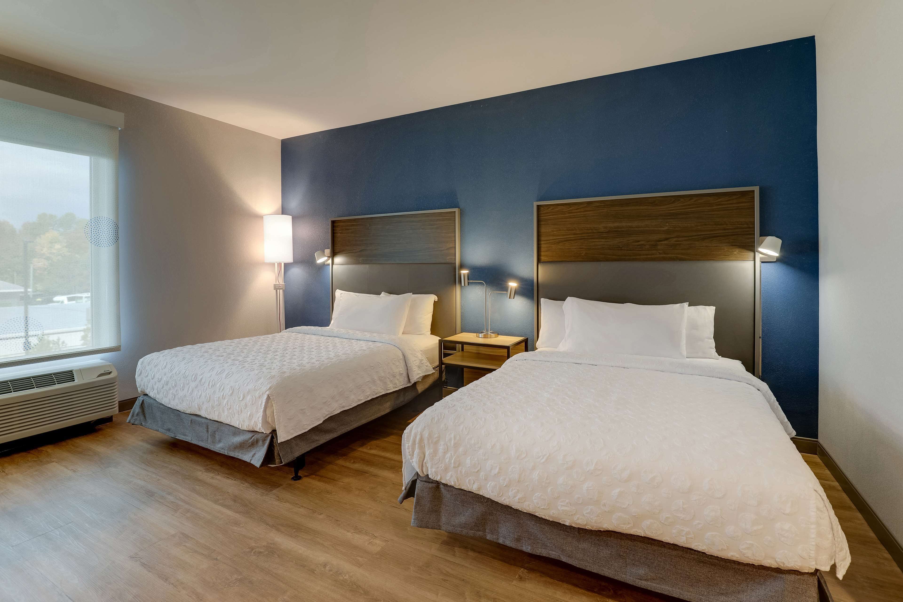 A hotel room with two beds, a window, and a wall-mounted air conditioner.
