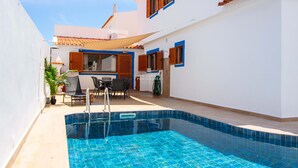 En swimmingpool med kakelbeklädd kant, en uteplats med utemöbler och en byggnad med blå fönsterramar och ett kaklat tak.