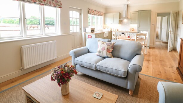 A spacious living room with a light blue sofa, a wooden coffee table with a vase of flowers, and large windows with floral curtains. - Flagstone Cottage (Cheltenham)