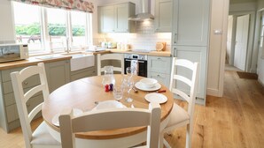 A modern kitchen with a dining area, featuring a wooden table, white chairs, and a view of the outdoors through large windows. - Flagstone Cottage (Cheltenham)