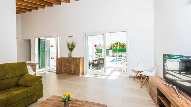 A modern living room with a green sofa, wooden coffee table, and a TV on a wooden stand. There is a sliding glass door leading to an outdoor area with a pool and furniture.