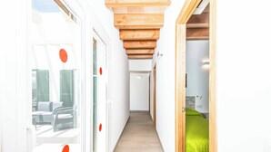 A narrow hallway with wooden ceiling beams, a bed with a green bedspread, and a view of outdoor furniture through a glass door.