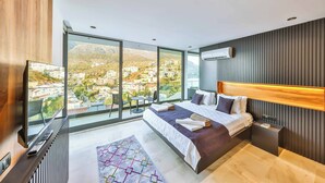 A modern bedroom with a large bed, a TV, and a view of a cityscape through large windows. - Villa Bank Two - Three Bedroom Villa, Sleeps 8 (Kalkan)