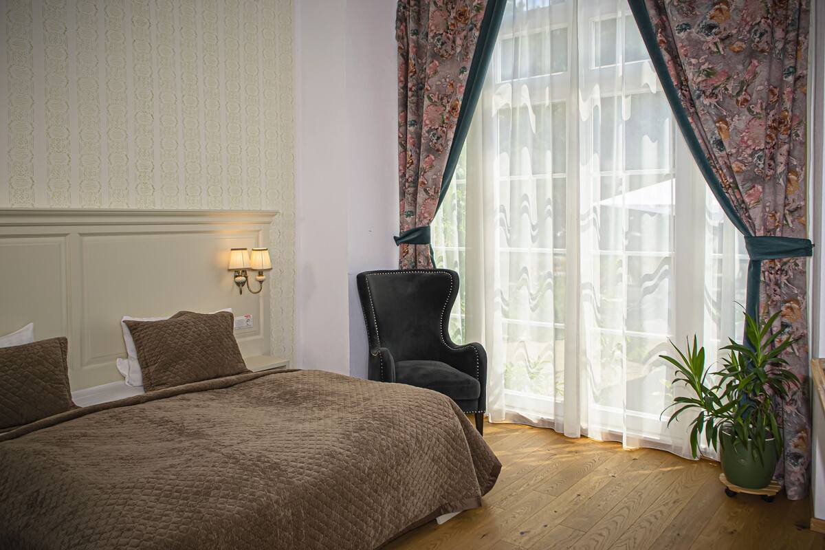 A bedroom with a large bed, a chair, a potted plant, and a window with curtains.