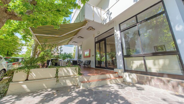 A covered outdoor seating area with a patio, a car parked nearby, and a building with large windows. - Hotel Avana Mare (Rimini)