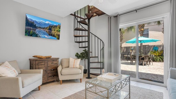 A modern living room with a spiral staircase, a sofa, a coffee table, and a large window overlooking a patio area. - 2 min walk to the beach from our Ocean Escape Jax Beach 3BR home! (Jacksonville Beach)