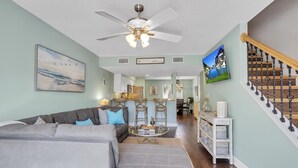 A living room with a grey sectional sofa, a ceiling fan, and a staircase with a wooden railing. - 2 min walk to the beach from our Ocean Escape Jax Beach 3BR home! (Jacksonville Beach)