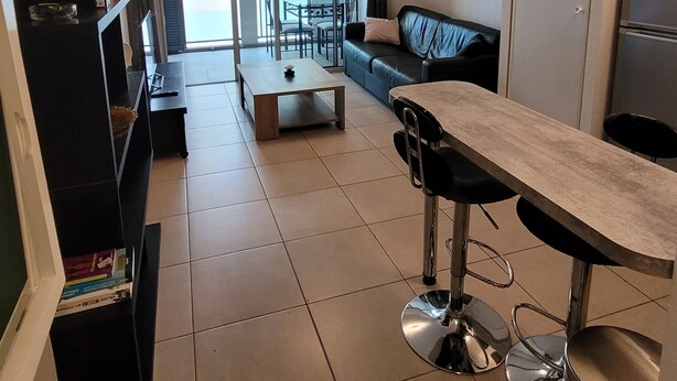 Un appartement moderne comprenant un espace de vie, une cuisine et un coin repas. On y trouve un canapé, une table basse, une table à manger avec des tabourets de bar et une bibliothèque.