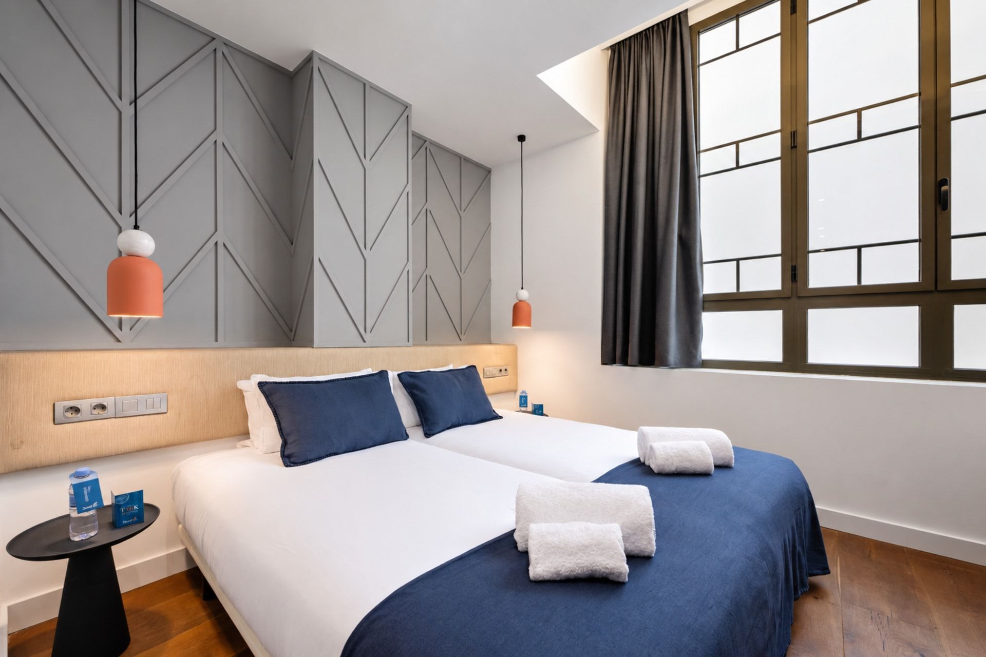 A hotel room with a bed, bedside tables, a window with curtains, and a wall with geometric patterns.