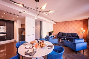 A modern living room with a blue sofa, a dining area with a round table, and a kitchen with a built-in oven.