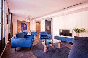 A modern living room with blue furniture, a flat-screen TV, and a dining area with a wooden accent wall.