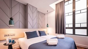 Ein modernes Hotelzimmer mit einem großen Bett, zwei Nachttischen, einem Beistelltisch, einem großen Fenster mit Vorhängen und geometrischen Wandverkleidungen.