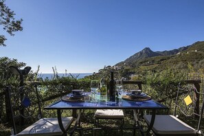 An outdoor dining area with a table set for two, overlooking a mountainous landscape and the sea.