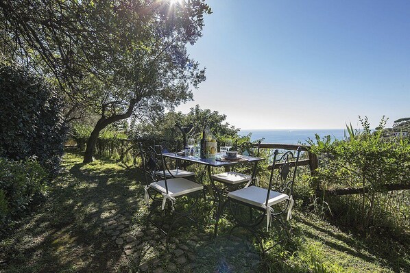 Un área de comedor al aire libre con mesa y sillas, rodeada de vegetación y con vista a un cuerpo de agua.