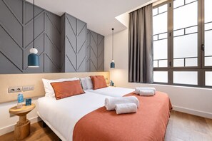 Een hotelkamer met een tweepersoonsbed, twee nachtkastjes, een houten nachtkastje, een wand met geometrische patronen, een raam met gordijnen en een hangende hanglamp.