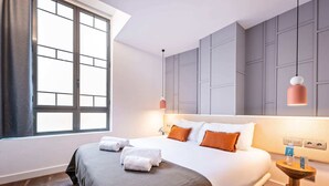 Habitación de hotel con una cama grande, paredes grises, una ventana con persianas y una mesita de noche con lámpara.