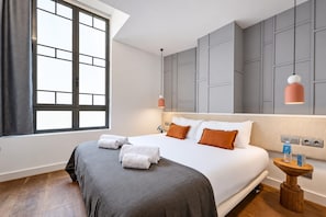 Une chambre d’hôtel avec un grand lit, une literie grise, un plancher en bois, une fenêtre avec des rideaux et une table de chevet avec une lampe.