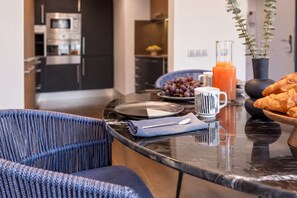 Une cuisine moderne avec un plan de travail en marbre, un coin repas agrémenté d’une chaise bleue, et un assortiment de produits pour le petit-déjeuner, notamment des croissants et un verre de jus d’orange.